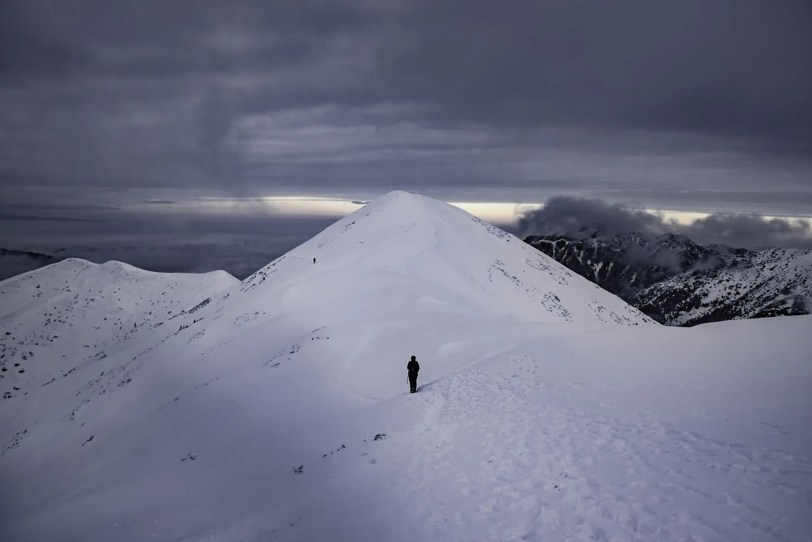 Hikers following a snow-covered mountain ridge beneath a cloudy sky.