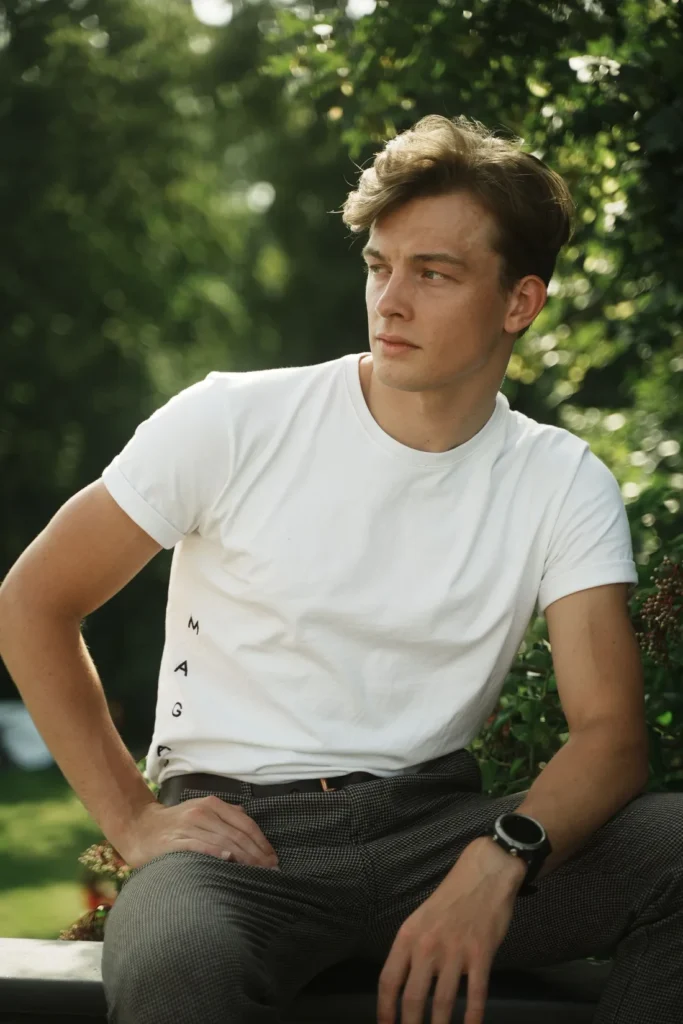 Man wearing a white short-sleeve T-shirt while sitting outdoors among green trees.